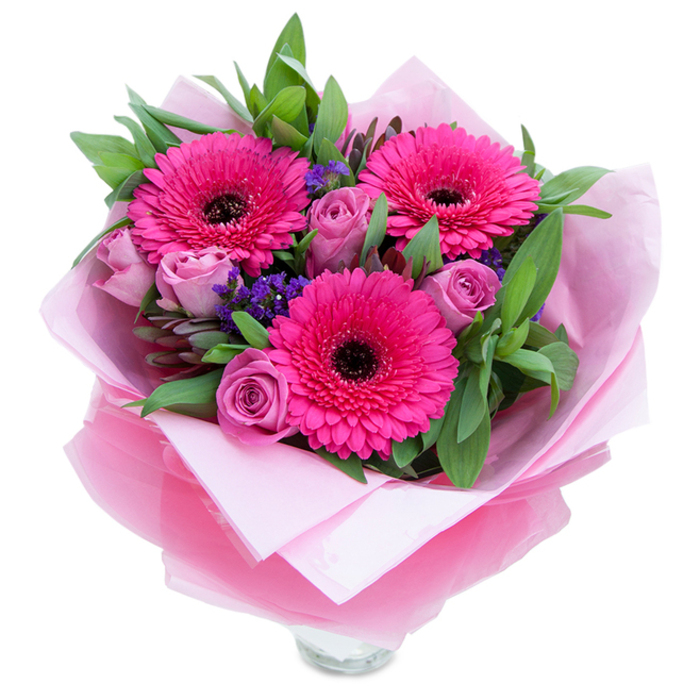 I think of this arrangement as a private, tender celebration in pinks, dominated by a trio of fuchsia Gerbera daisies that open wide like joyful faces, each with a dark, defining centre that anchors the composition. Surrounding those energetic blooms are roses in a muted lavender-pink or dusty rose, their petals still closely furled in the soft geometry of youth-petals that look and feel velvety under the fingertips and carry a delicate rose scent. Interspersed are tiny purple-blue sprigs of statice or limonium that float between the roses and gerberas, adding texture and a hint of countryside whimsy. The green foliage-broad, glossy leaves reminiscent of salal or eucalyptus-forms a generous backdrop that makes the pinks sing and suggests freshly gathered stems. The bouquet is thoughtfully presented: an inner wrap of pale pink tissue paper provides a soft, matte base while an outer layer of translucent fuchsia-pink cellophane, faintly crinkled, introduces a gentle shimmer and reinforces the pink theme; the wrapping is folded with care so the flowers sit snug and secure. At the base you can see the top rim of a clear glass vase, indicating immediate display and the freshness of water-cut blooms. Under soft daylight the petals reveal subtle tonal shifts, the roses exuding a faint perfume, gerberas lending a green-sweet hint, and the statice offering a dry, airy contrast. This composition feels right for placing on a bedside table, sending to a loved one in Ruislip Manor or popping into a Ruislip High Street flat to mark an anniversary, birthday or simply to say 'I'm thinking of you.'